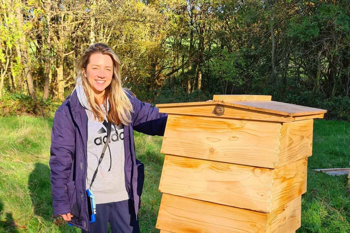 A person with blond hair smiling next to a large wooden beehive in a sunny, green outdoor setting.
